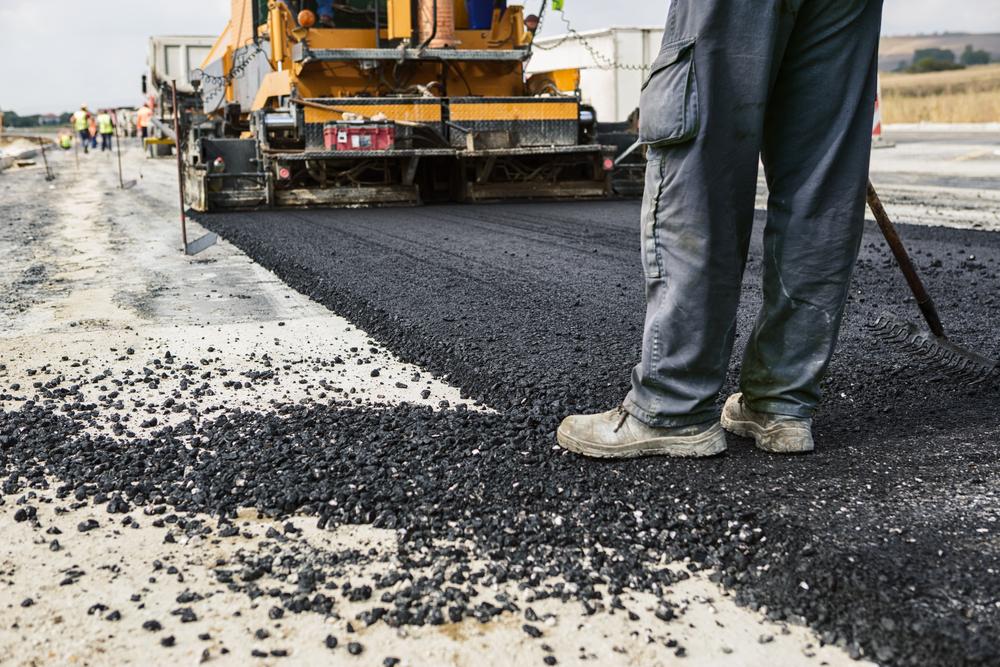 Legs of a worker standing on asphalt with a paving machine in the background